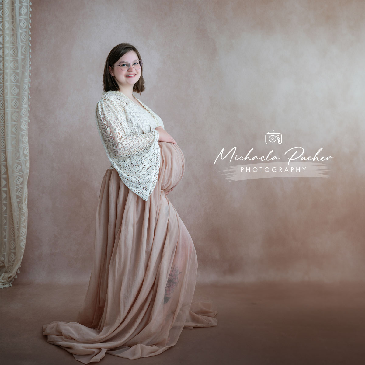 Hochschwangere Frau im Studio steht seitlich vor einem hellen Hintergrund, trägt eine cremefarbene Spitzenjacke und einen fließenden roséfarbenen Stoffrock und hält sanft ihren Babybauch.